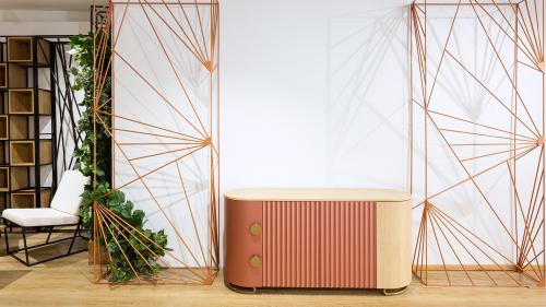 The image showcases a compact T-DESIGN sideboard featuring a distinctive copper-blush front that brings warmth and elegance to the space. The finely milled fluted surface is complemented by natural oak accents and brass details, creating a timeless yet contemporary piece of furniture.  The sideboard is framed by decorative REZES BANDA room dividers, which serve not only as functional space separators but also as striking design elements that define the interior.
